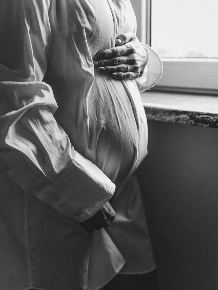 Femme enceinte en chemise blanche près d’une fenêtre, les mains posées sur son ventre